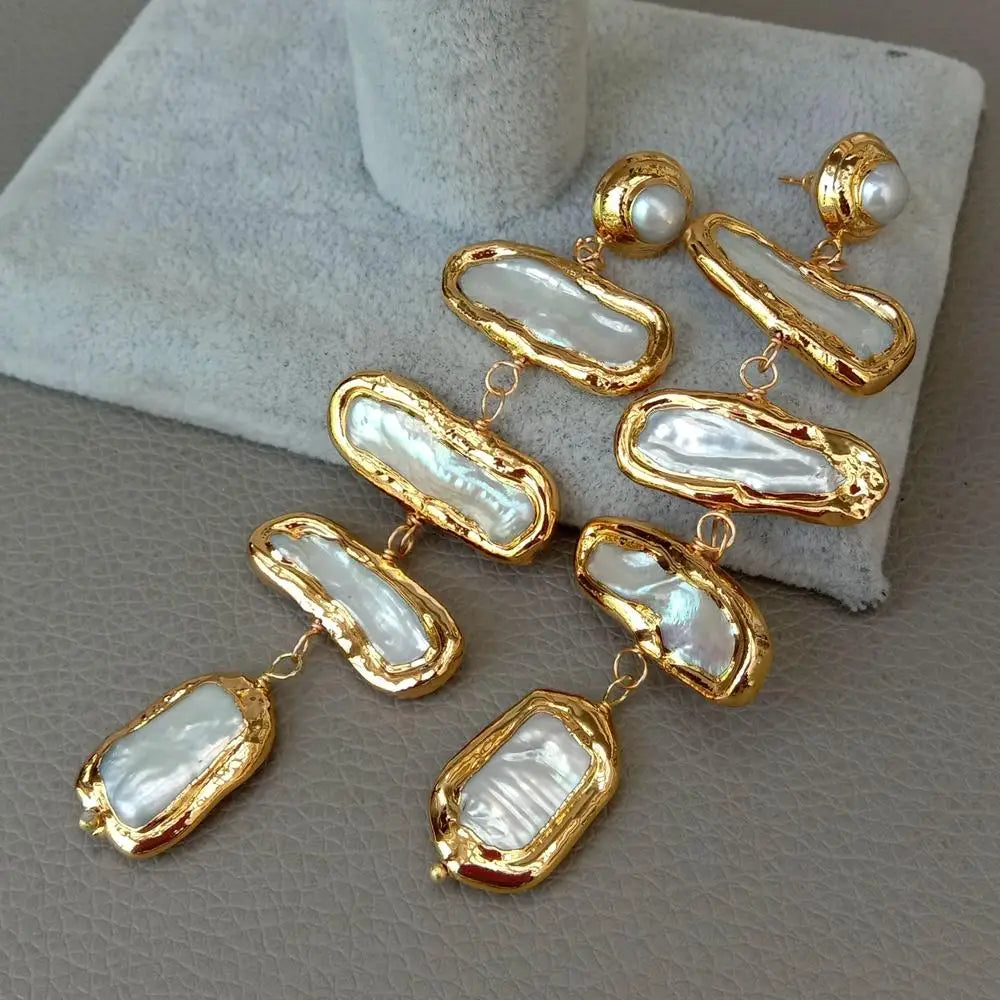 A pair of gold-tone drop earrings with irregular white pearl-like stone segments and small pearl studs at the top, displayed on a light gray jewelry stand against a gray textured surface.