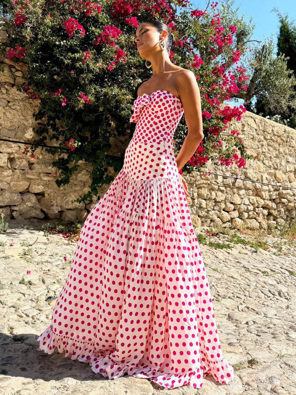Woman in a pink polka dot dress standing in front of a stone wall and flowering bush.