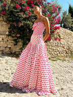 Woman in a pink polka dot dress standing in front of a stone wall and flowering bush.