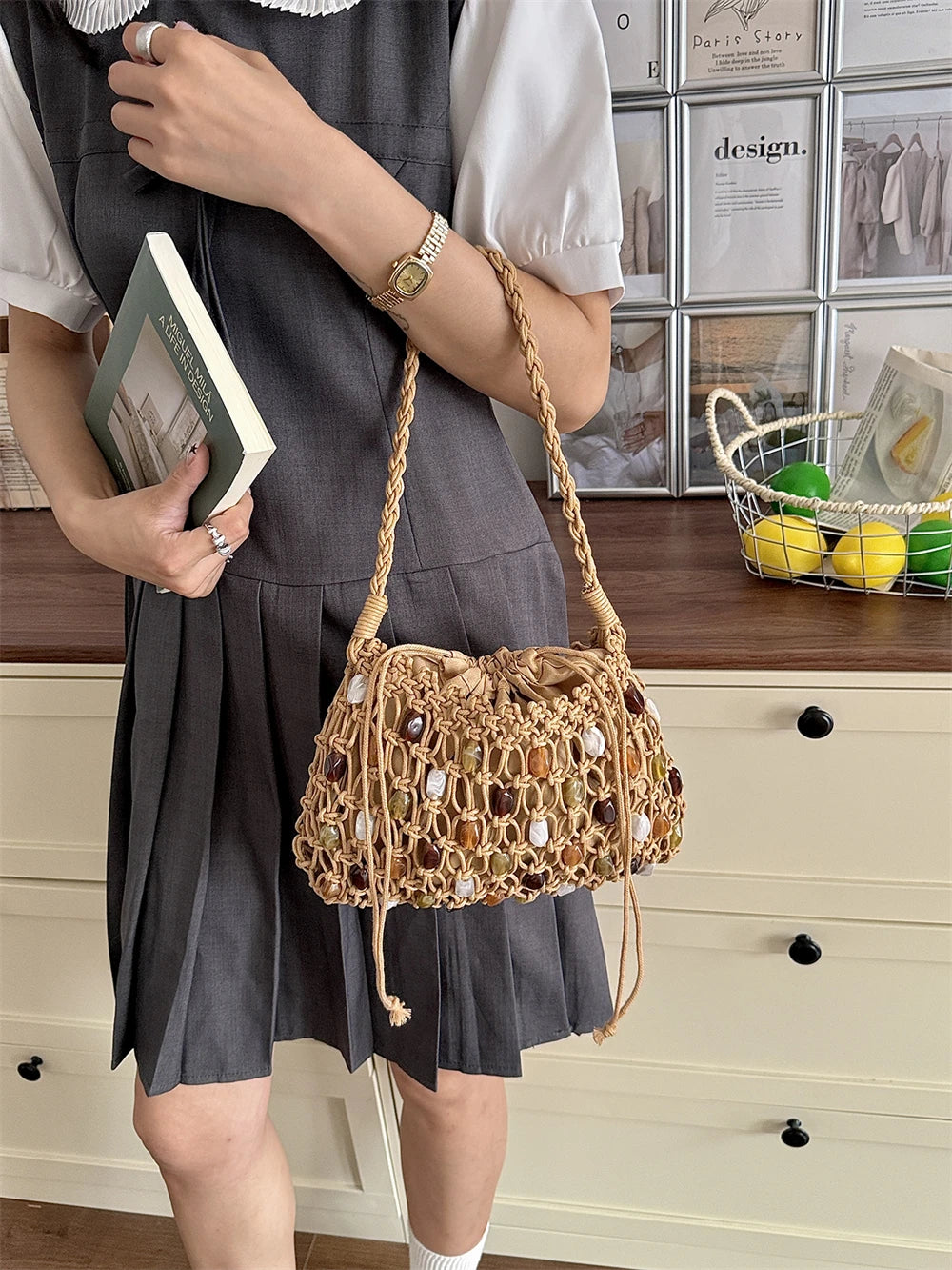 A person wearing a gray pleated dress and white short-sleeve blouse holds a woven straw shoulder bag with a braided strap in front of a cream-colored dresser and framed wall art.