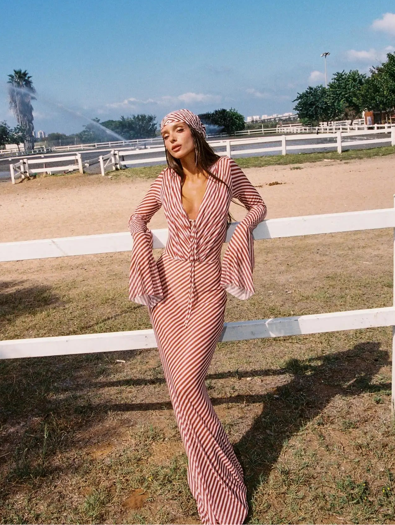Woman in a pink dress standing in an outdoor setting with a white fence and trees in the background.