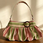 A pleated shoulder bag with vertical stripes in sage green, dusty pink, and cream, featuring a wide matching strap and a prominent round brown metal ring detail on the front, resting on a wooden table.