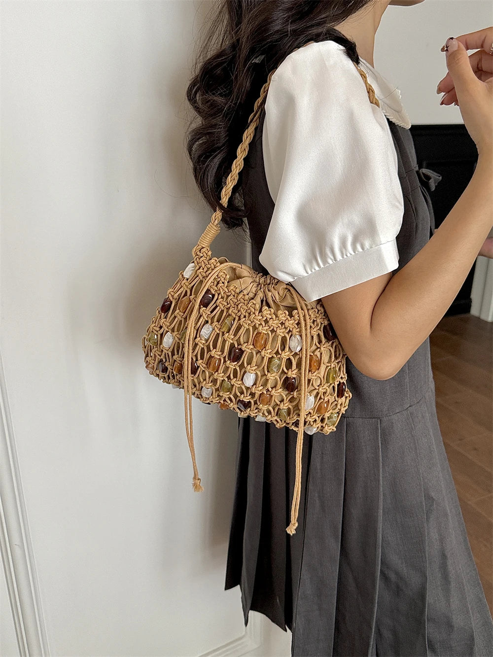 A woman wearing a white puff-sleeve top and a dark gray dress carries a tan crochet bucket bag with a braided shoulder strap and drawstring closure.