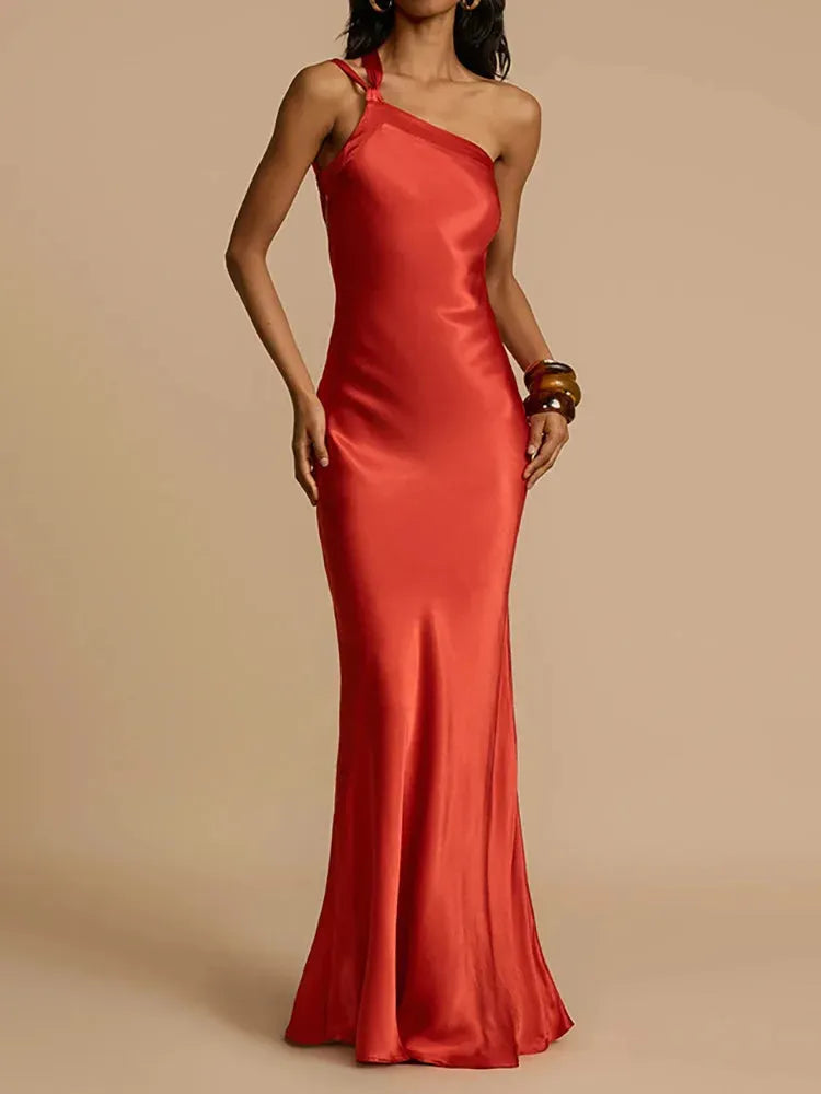 A woman wearing a red, solid color, diagonal collar, spaghetti strap, maxi dress that reaches the floor.