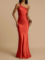 A woman wearing a red, solid color, diagonal collar, spaghetti strap, maxi dress that reaches the floor.