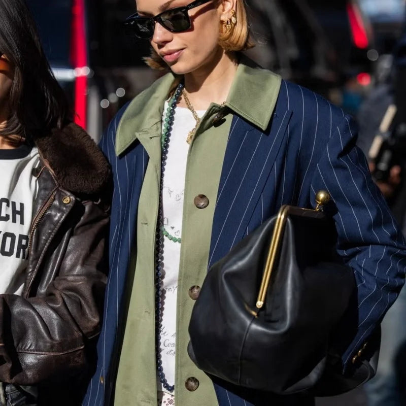 A person wearing a navy pinstripe coat over an olive green shirt, carrying a large black leather handbag with gold hardware in an outdoor street setting.