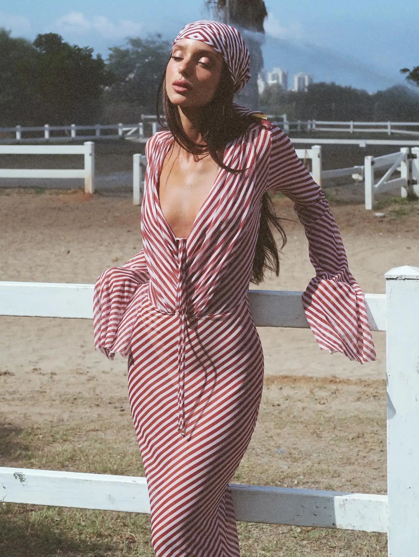 Woman wearing a red and white striped dress in an outdoor setting with a white fence and trees in the background.