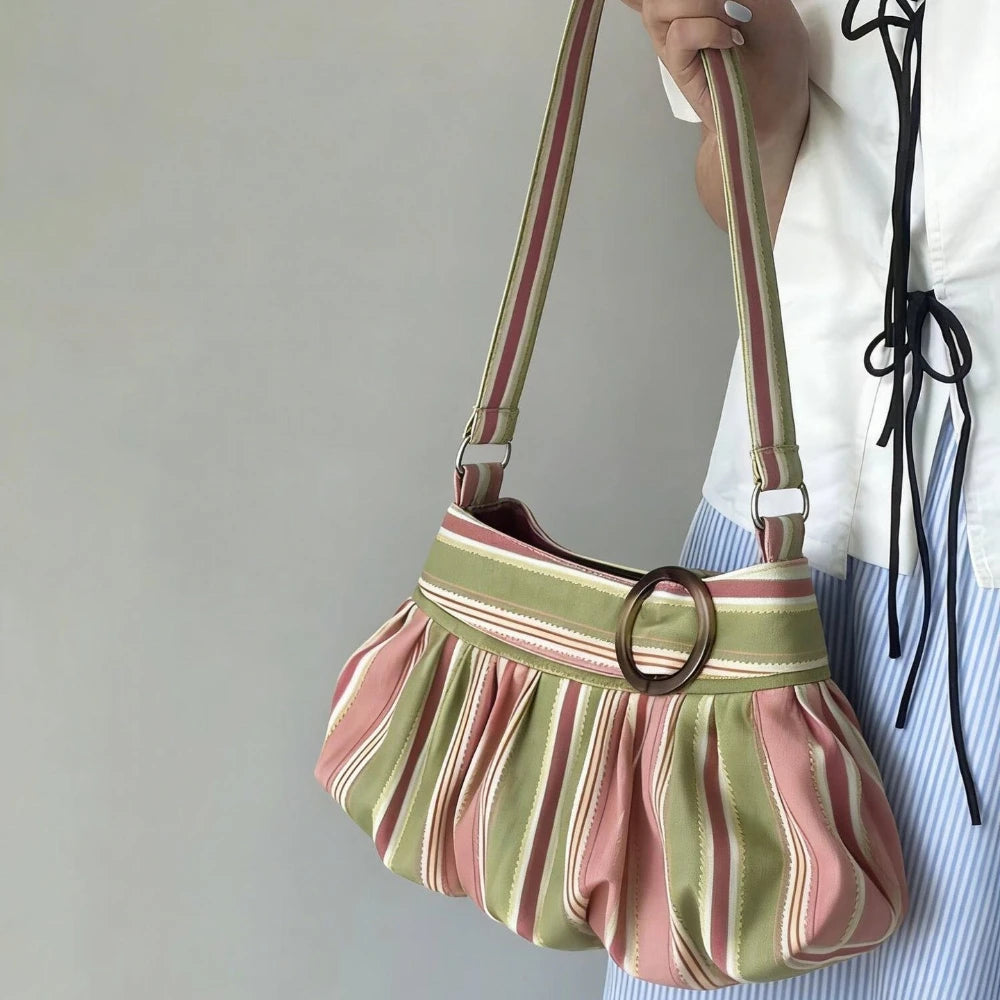 A person holds a pleated shoulder bag with vertical stripes in sage green, dusty pink, and cream. The bag features a wide top band with a central dark brown wooden ring and a matching striped strap.