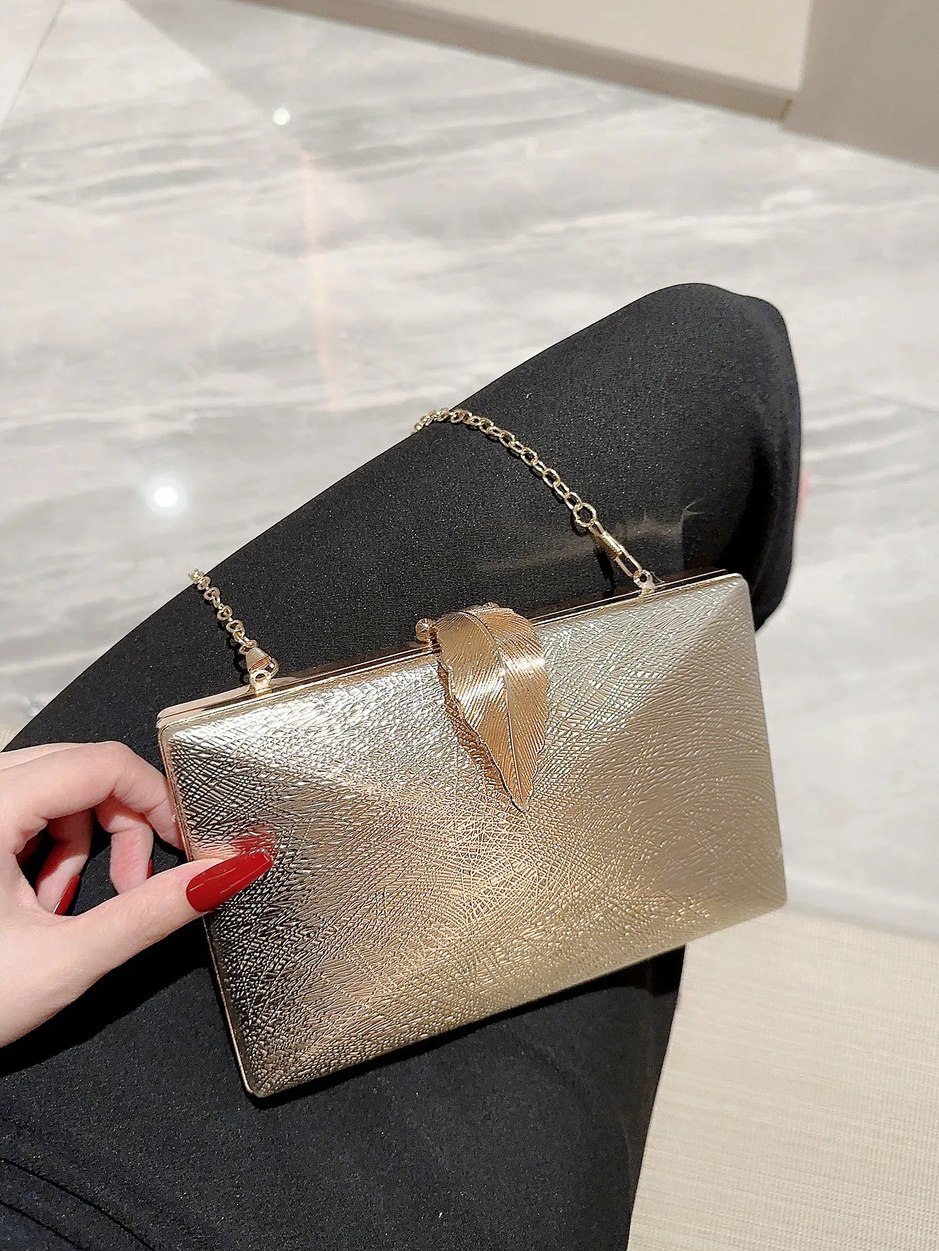 Gold clutch bag held by a hand with red nail polish on a marble surface
