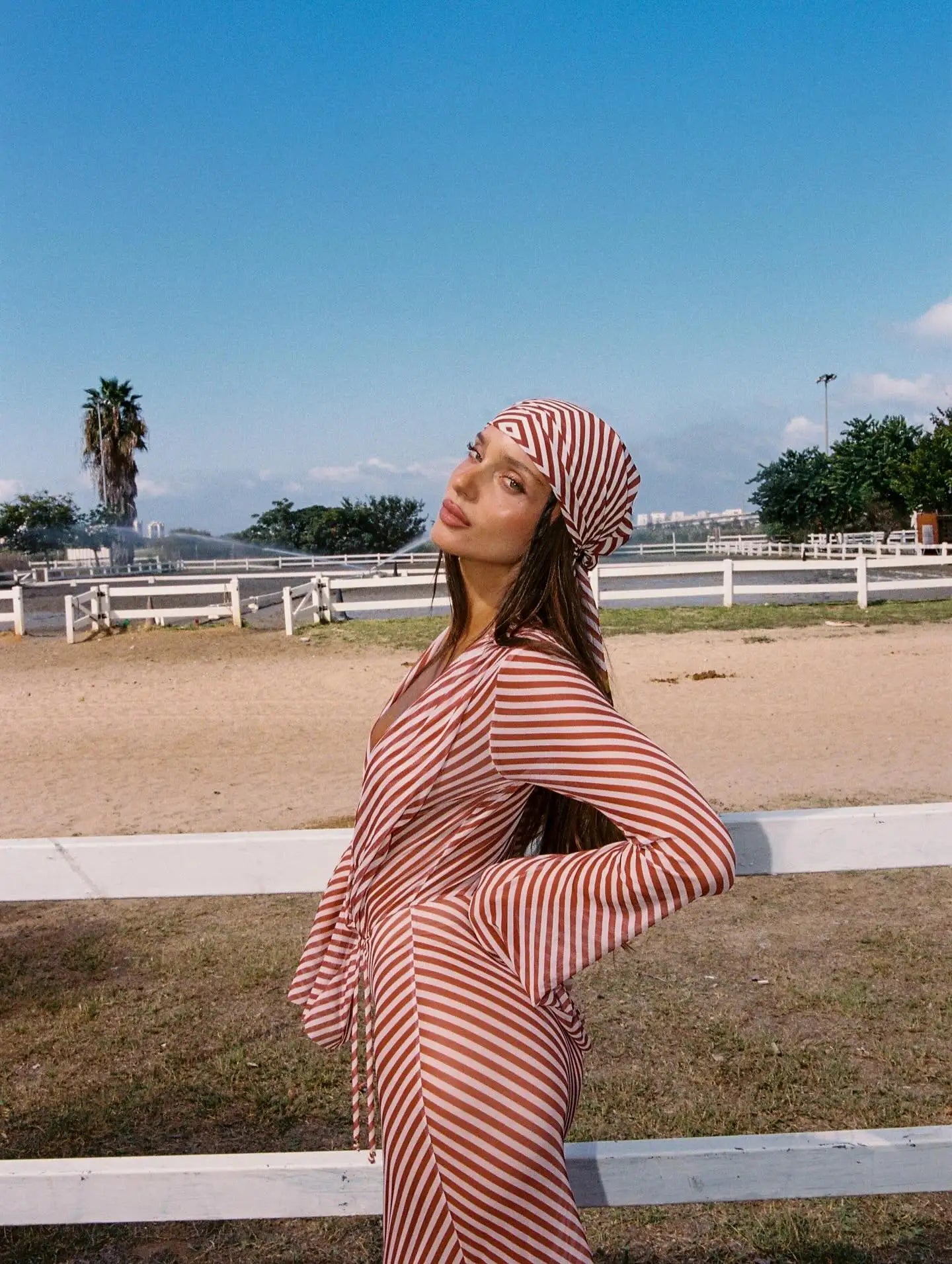 Woman in a red and white striped dress and headscarf standing outdoors with a clear blue sky.