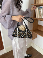 A person in a light lavender blouse and white skirt holds a black crochet bucket bag with a cream drawstring and multicolored stone-like accents, standing indoors near a wooden bookshelf.