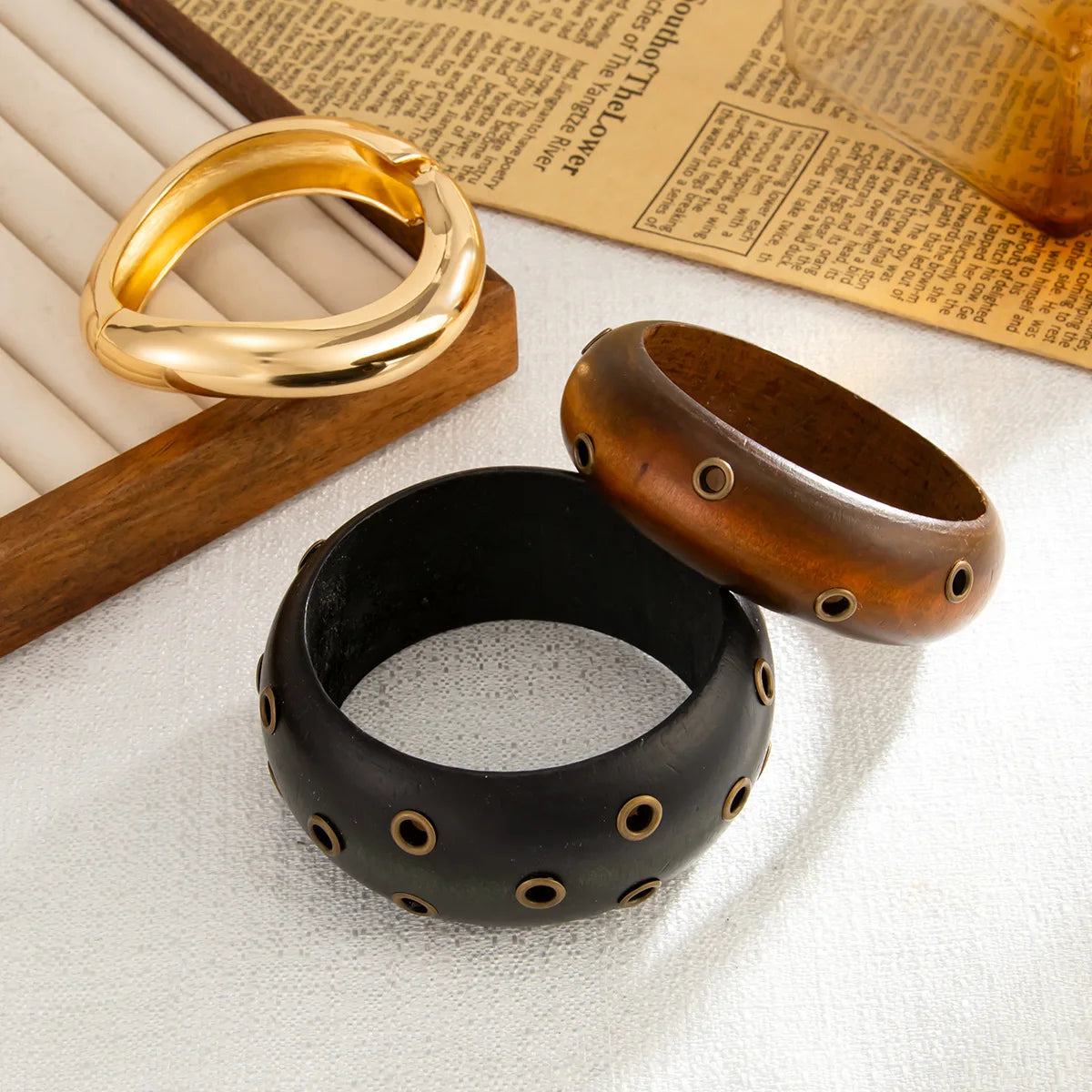 Three bracelets in gold, black, and brown on a textured surface with a newspaper in the background.