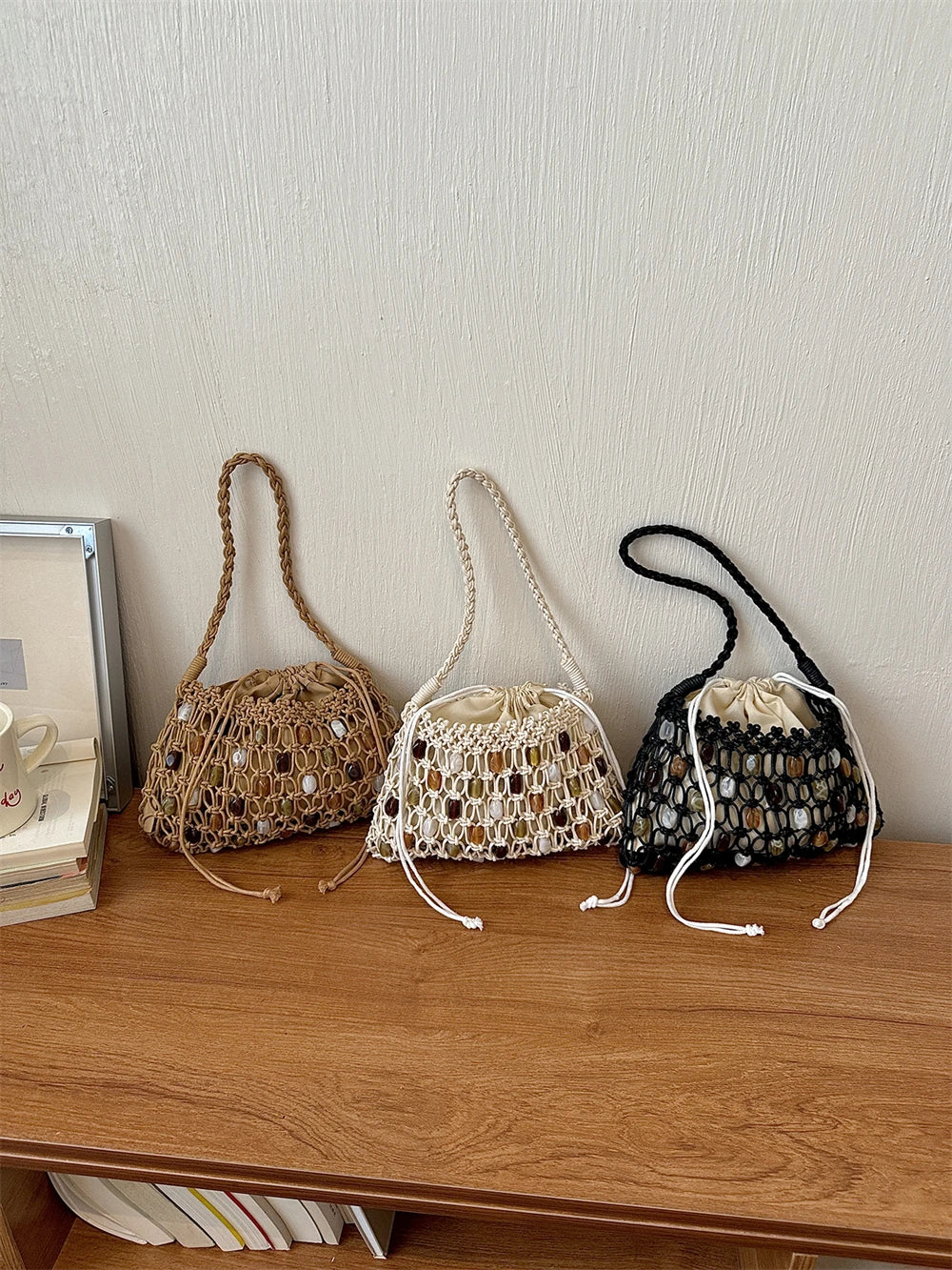 Three crocheted bucket bags with woven stone accents are lined up on a wooden shelf against a textured off-white wall. The bags are in natural tan, cream, and black, each with a braided shoulder strap and drawstring closure.