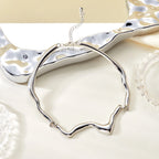 A polished silver-toned open bangle necklace with a wavy, organic silhouette and a short extender chain, displayed on a light cream surface with a reflective silver prop in the background.