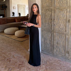 A woman stands indoors wearing a sleeveless black maxi dress with gold embellishments along the shoulder straps. She is holding a small black handbag and standing in a luxurious room with a large carved wooden cabinet, a terracotta tile floor, and a sofa in the background.