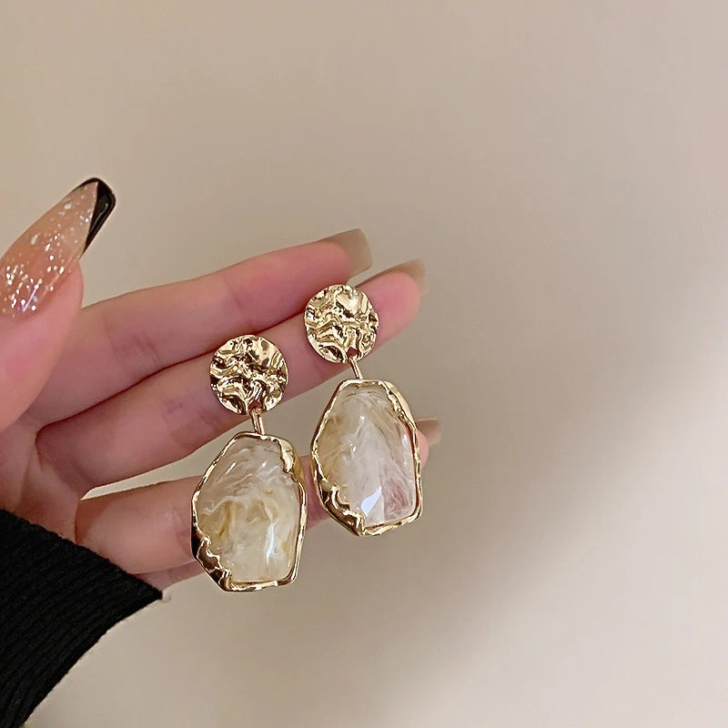 Gold earrings with large stones held by a hand against a neutral background