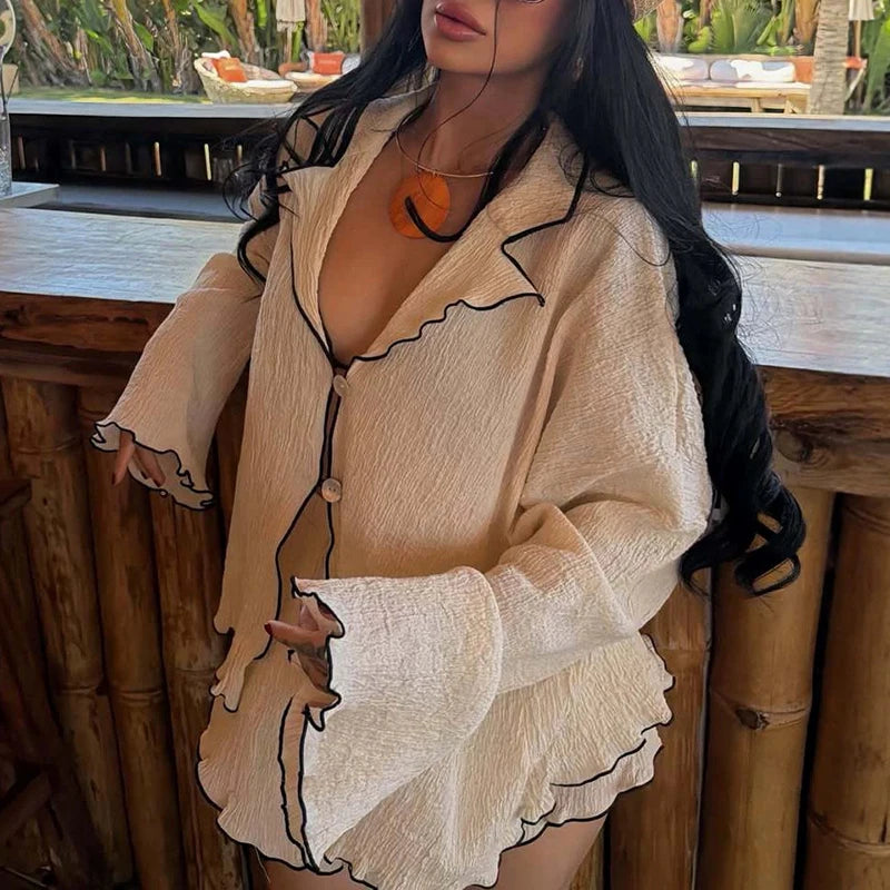 A woman wearing a beige crinkled long-sleeve pajama-style top with black piping and a deep V-neck, photographed in a real bar or lounge setting with wooden railings and plants in the background.
