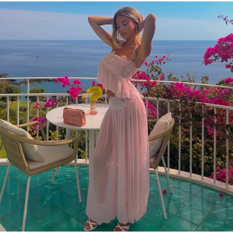 A woman stands on a balcony overlooking the ocean, wearing a light pink two-piece maxi dress with a ruffled crop top and a flowing skirt. The setting includes a white table and chairs, bright pink bougainvillea flowers, and a turquoise tiled floor.