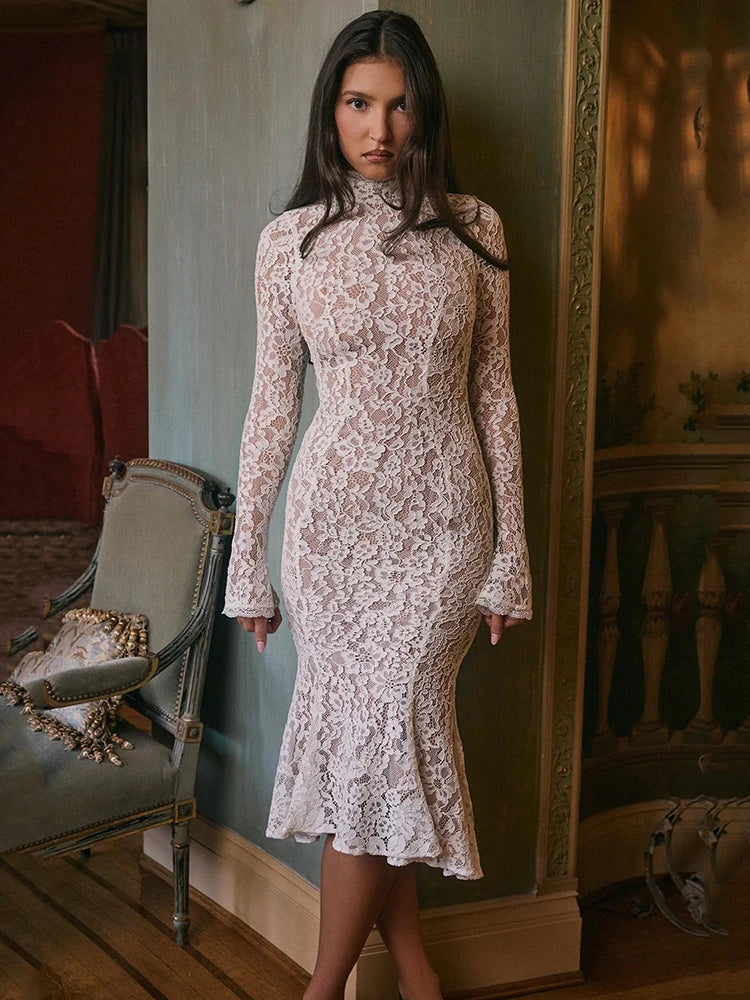 Woman wearing a lace dress standing in an elegant room with a mirror and chair.