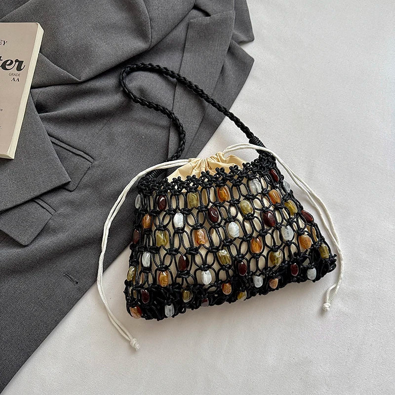 A black crocheted drawstring bucket bag with woven multicolor stone accents, styled on a white surface next to a gray blazer and a book corner.