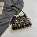 A black crocheted drawstring bucket bag with woven multicolor stone accents, styled on a white surface next to a gray blazer and a book corner.