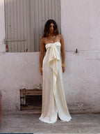 Woman wearing a strapless white gown with a bow detail against a plain wall.