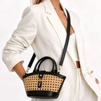 A woman in a white blazer carries a small structured handbag with black leather trim and handles, plus a natural woven cane or raffia front panel and a black shoulder strap.