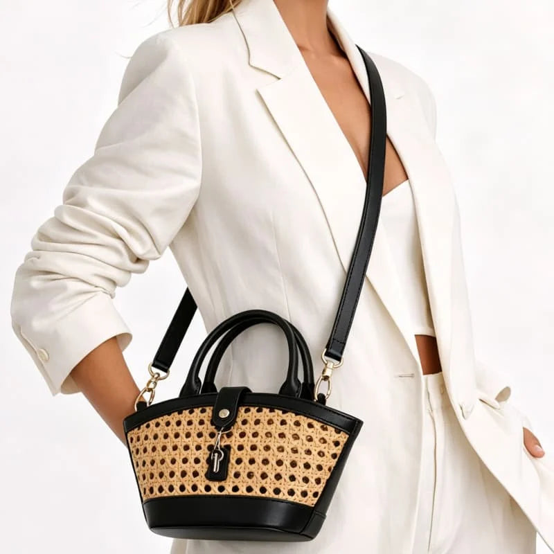 A woman in a white blazer carries a small structured handbag with black leather trim and handles, plus a natural woven cane or raffia front panel and a black shoulder strap.