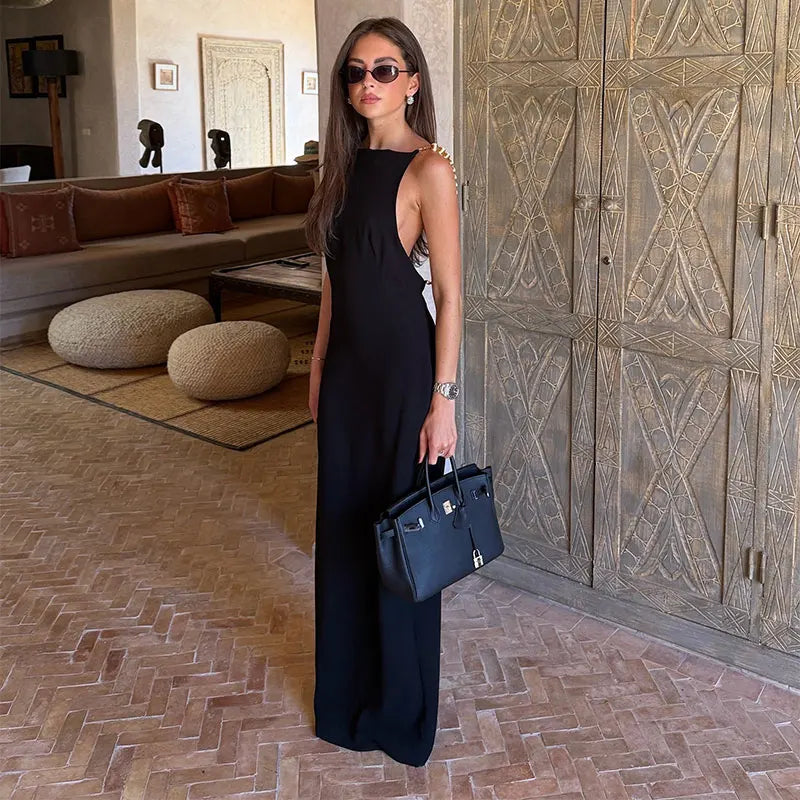 A woman wearing a sleeveless black maxi dress and sunglasses stands indoors holding a structured black handbag with gold-tone hardware, set against a luxury interior with patterned wooden doors and a herringbone tile floor.