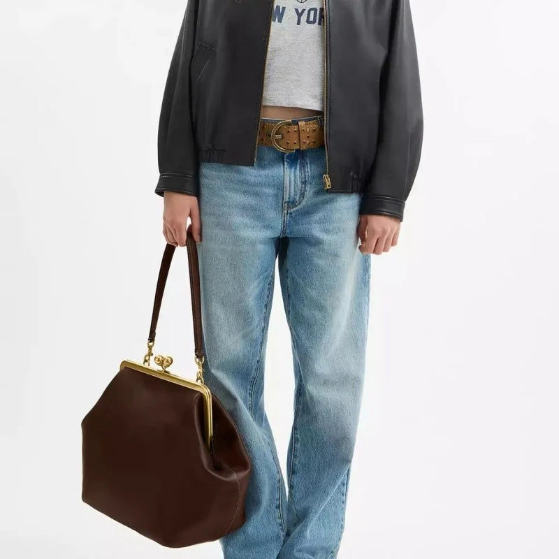 A person wearing a black leather bomber jacket, gray graphic t-shirt, and light-wash blue jeans, holding a dark brown leather kiss-lock handbag with gold hardware against a plain white background.