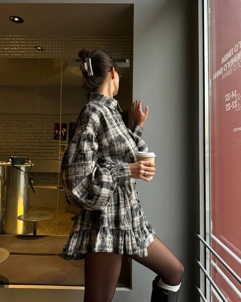 A woman stands in a cafe wearing a black and white plaid long-sleeve mini dress, holding a white paper coffee cup.