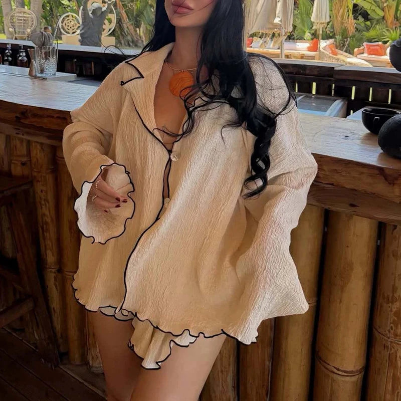 A woman wearing a beige crinkled long-sleeve cover-up with black trim over a bikini, standing in front of a bamboo-wood bar.