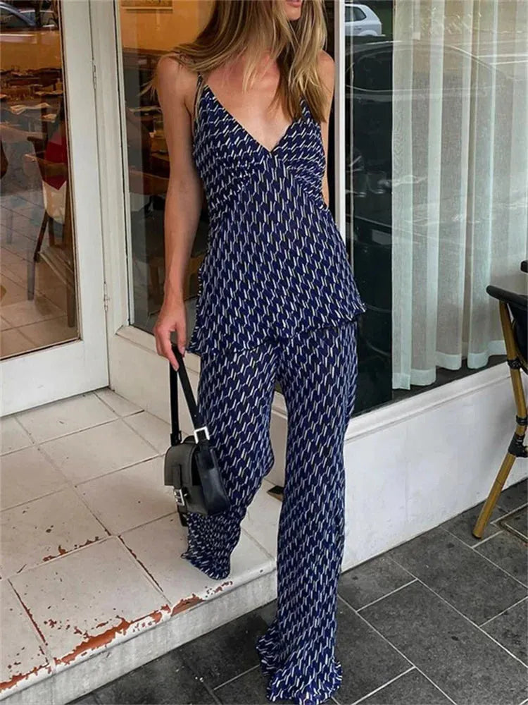 A woman wearing a navy two-piece outfit with a geometric pattern, consisting of a sleeveless top with a V-neck and wide-leg pants, standing in front of a glass door.