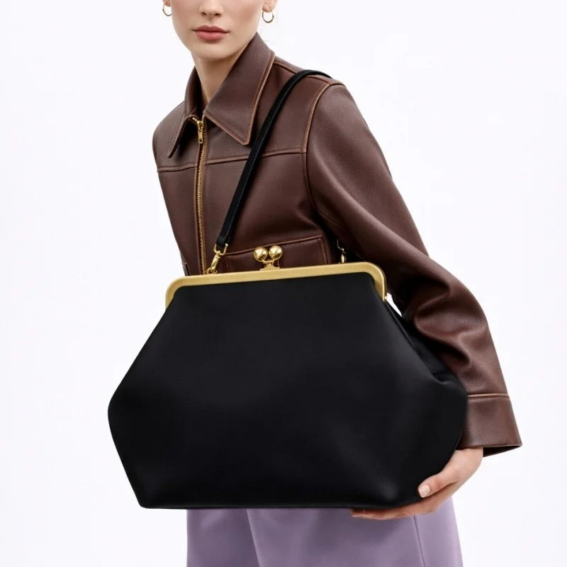 A model wearing a brown leather jacket and lavender pants carries a large black handbag with a gold-tone kiss-lock frame and shoulder strap against a plain white background.