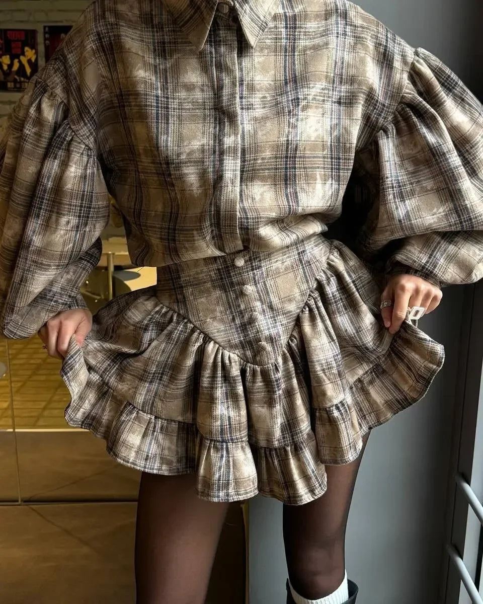 A person wearing a beige and brown plaid mini dress with dramatic puffed long sleeves, a button front, and a ruffled tiered skirt, paired with sheer black tights and white socks.