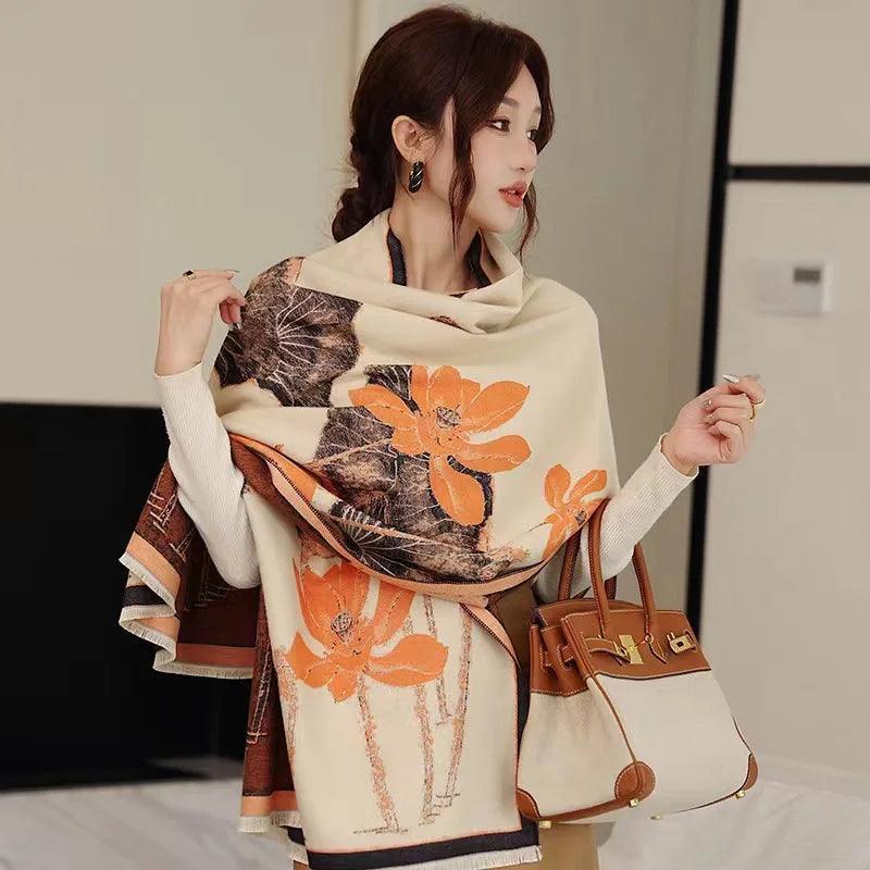 A woman wearing a beige, brown, and orange floral cashmerette scarf with a print pattern, paired with a white top and beige pants. She is also carrying a brown purse.