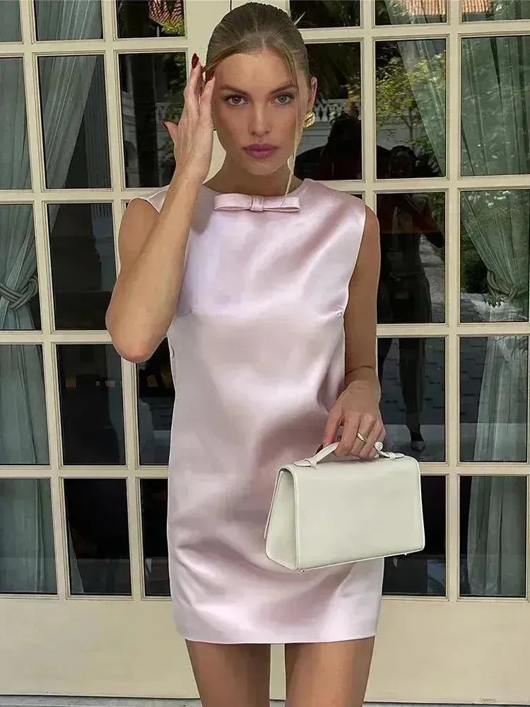 Woman in a light pink dress holding a white handbag in front of a window.