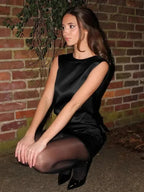 Woman in black dress sitting against a brick wall.