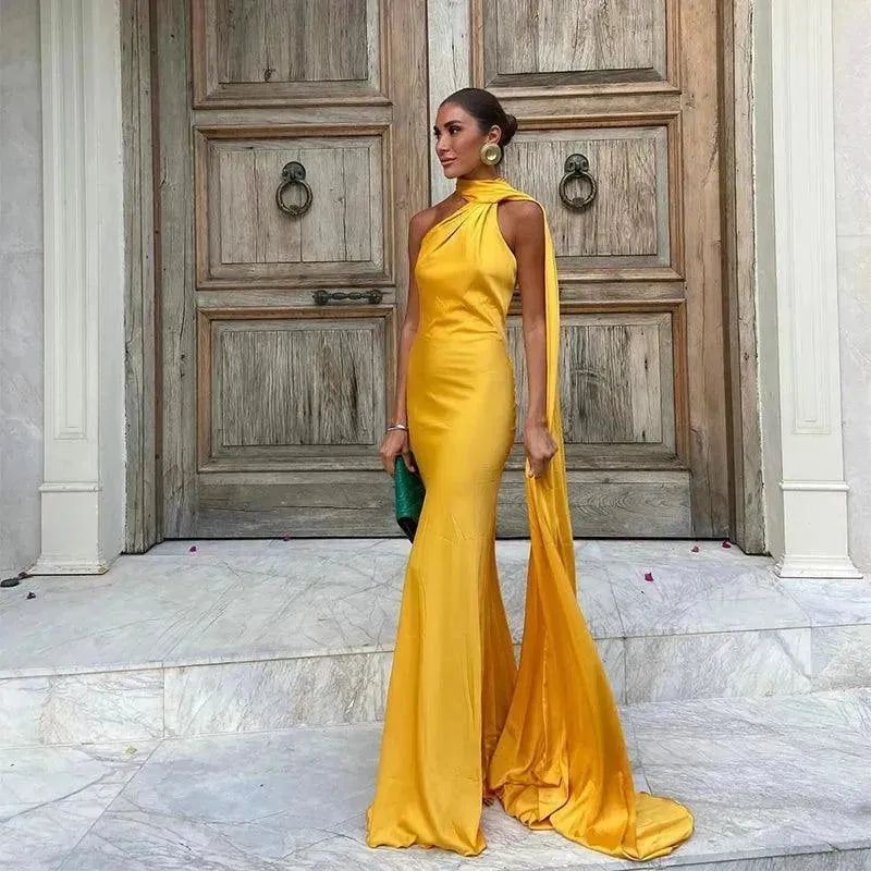 A woman standing on a staircase wearing a yellow maxi dress with a halter neckline and backless design, holding a green purse and wearing earrings.
