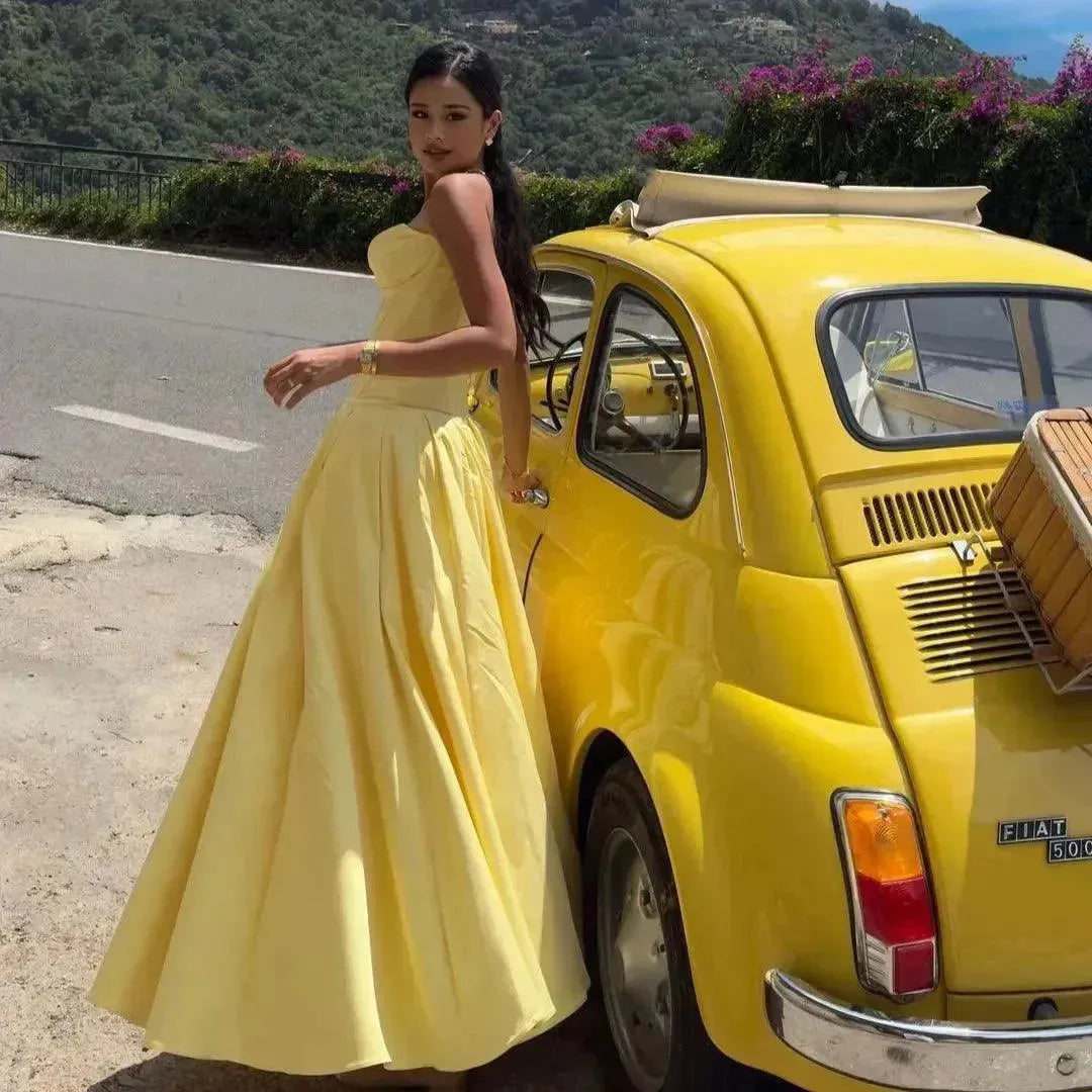 Woman in a yellow dress standing next to a yellow car on a mountain road.