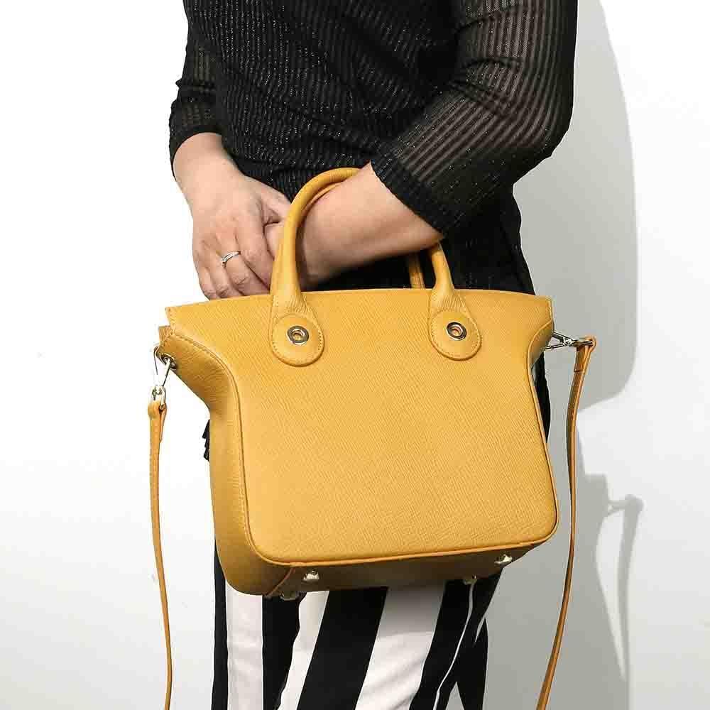 A person holding a yellow leather tote bag with a zipper and a cell phone pocket, featuring a trapezoidal shape and a single strap design.